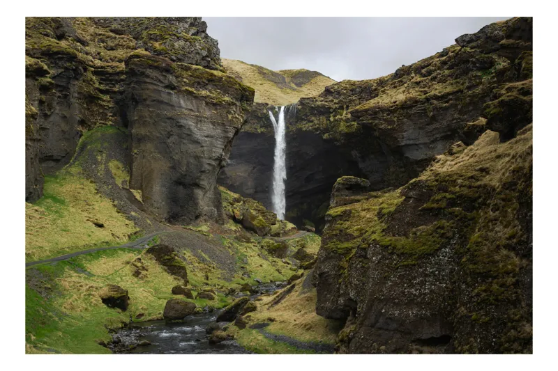 Tablou Canvas, "Cascada ascunsă între stânci verzi", Cascadă, Stânci, Râu, Verdeață, Cer Înnorat, Peisaj Montan, Natură, Frumusețe Sălbatică, Tranquilitate, Aventură, 100 x 70 cm