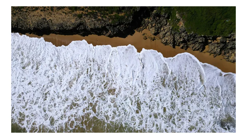 Tablou Canvas, "Valurile mării îmbrățișând țărmul stâncos", Spumă, Valuri, Plajă, Nisip, Stânci, Vegetație, Coastă, Ocean, Nature, Persepectivă, Aerian, Liniște, Contrast, Texturi, Albastru., 90 x 50 cm