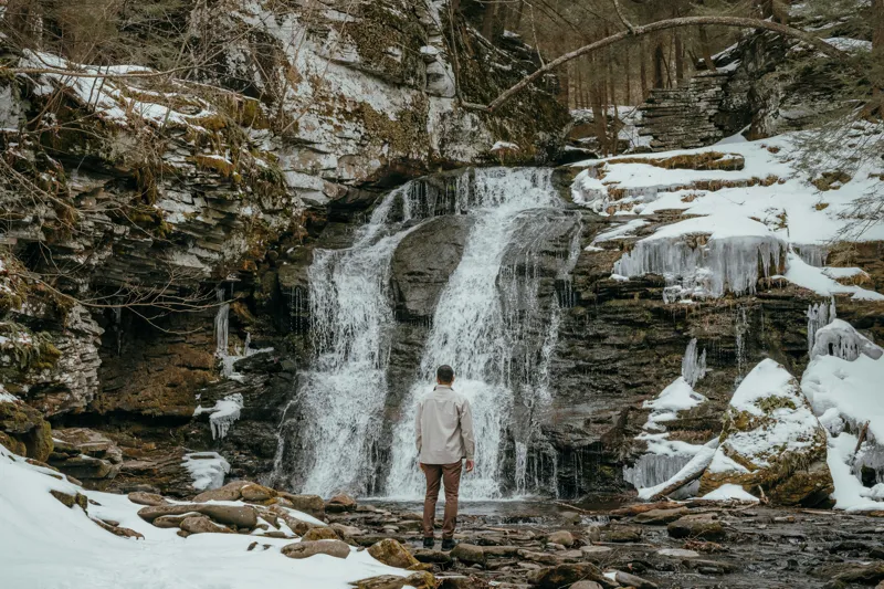 Tablou Canvas, "Om admira cascada înzăpezită iarna", Cascadă, Zăpadă, Gheață, Pădure, Roci, Figură Umană, Iarnă, Natură, Tranquilitate, Aventură, 150 x 100 cm – poza 2